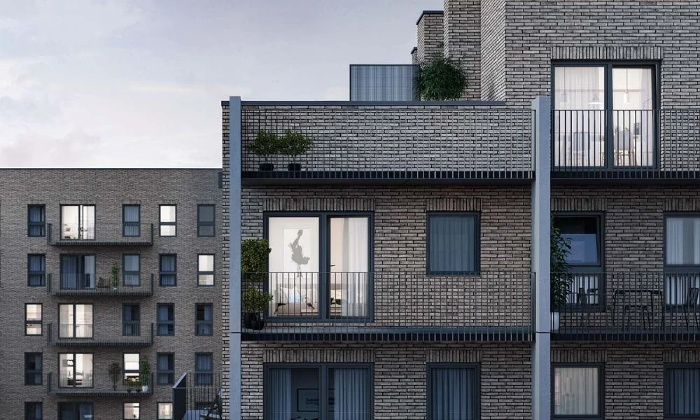 Mieszkaniowy Dzień Otwarty Nowoczesny budynek mieszkalny z balkonami i dużymi oknami, w jednym z mieszkań widoczna sylwetka osoby na tle drzwi balkonowych, elewacja z szarej cegły, rośliny w donicach na balkonach.