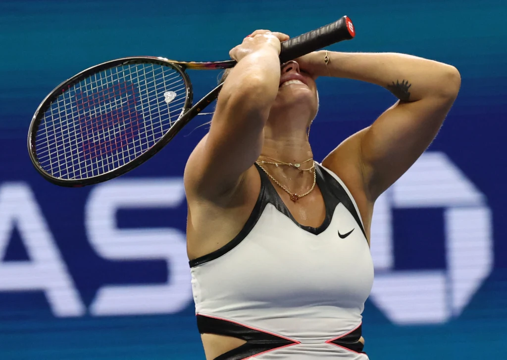 Zawodniczka tenisa w sportowym stroju trzyma rakietę nad głową i zakrywa twarz rękami, wyrażając silne emocje na korcie.