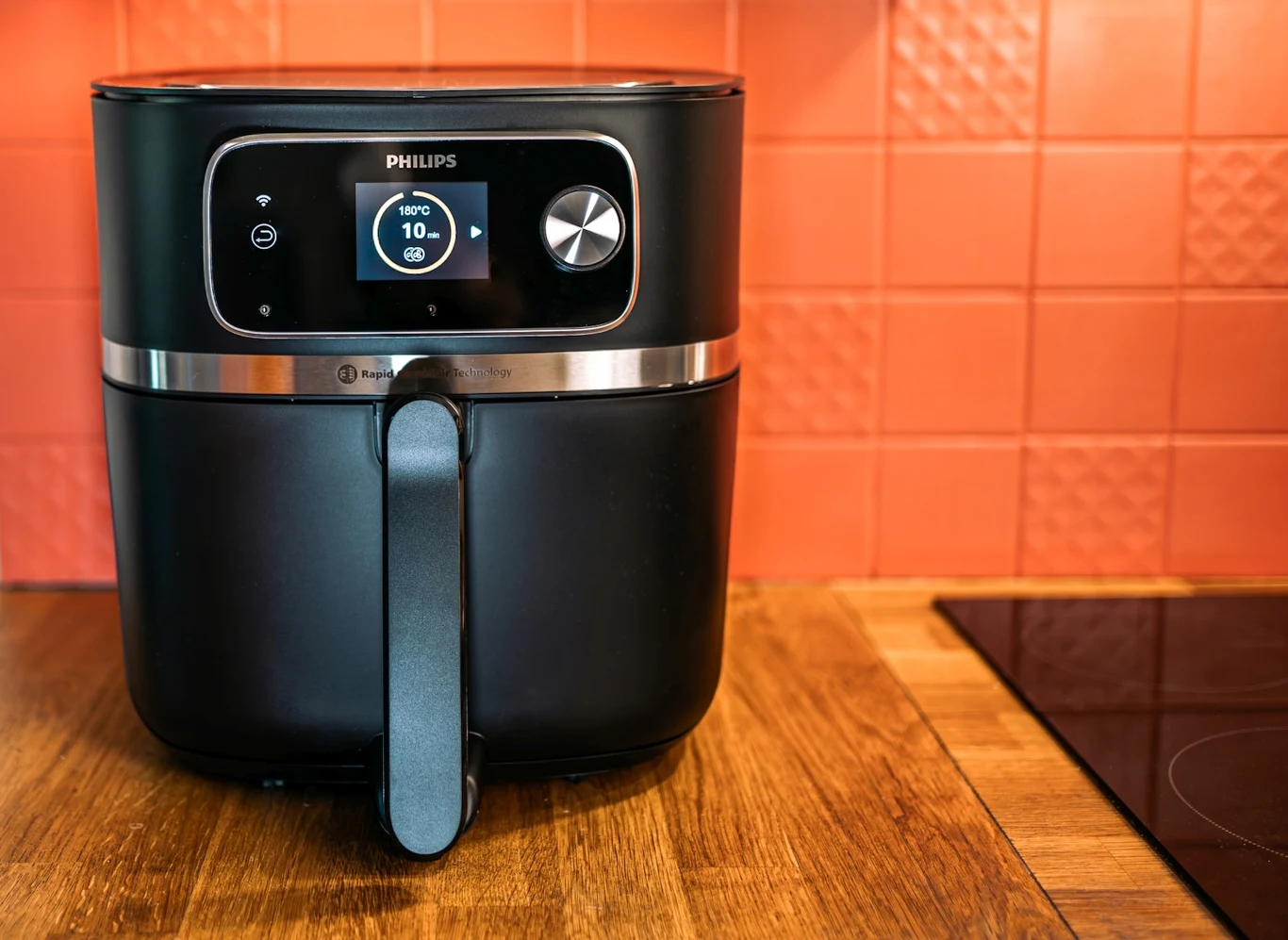 Potrawy przygotowywane w Airfryerze niemal nie wymagają użycia tłuszczu Czarna frytkownica beztłuszczowa marki Philips ustawiona na drewnianym blacie kuchennym na tle pomarańczowych płytek, obok widoczna część płyty grzewczej.