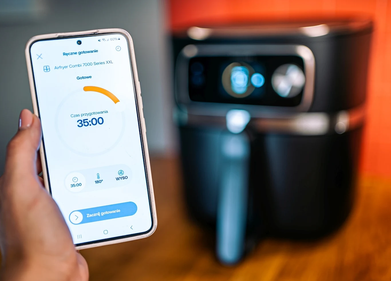 Aplikacja HomeID pozwala sterować urządzeniem z poziomu smartfona i zawiera mnóstwo przepisów Aplikacja na smartfonie pokazuje czas przygotowania potrawy w frytkownicy beztłuszczowej, urządzenie stoi na blacie kuchennym z lekko rozmytym tłem, dominuje motyw nowoczesnej technologii w kuchni.