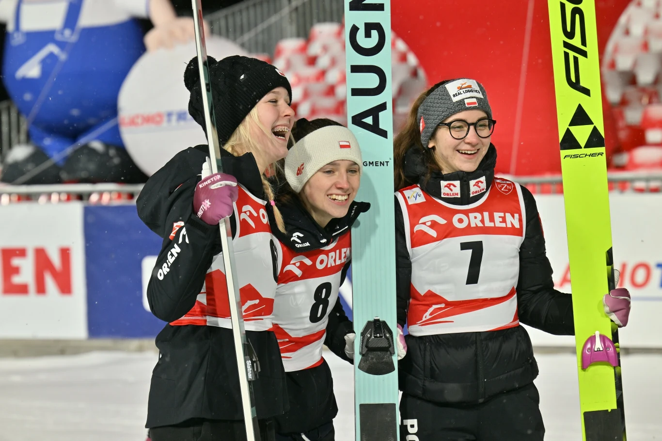 Trzy zawodniczki w sportowych strojach zimowych, ubrane w numery startowe z logo sponsora Orlen, śmieją się i celebrują razem, trzymając narty w otoczeniu sportowej areny