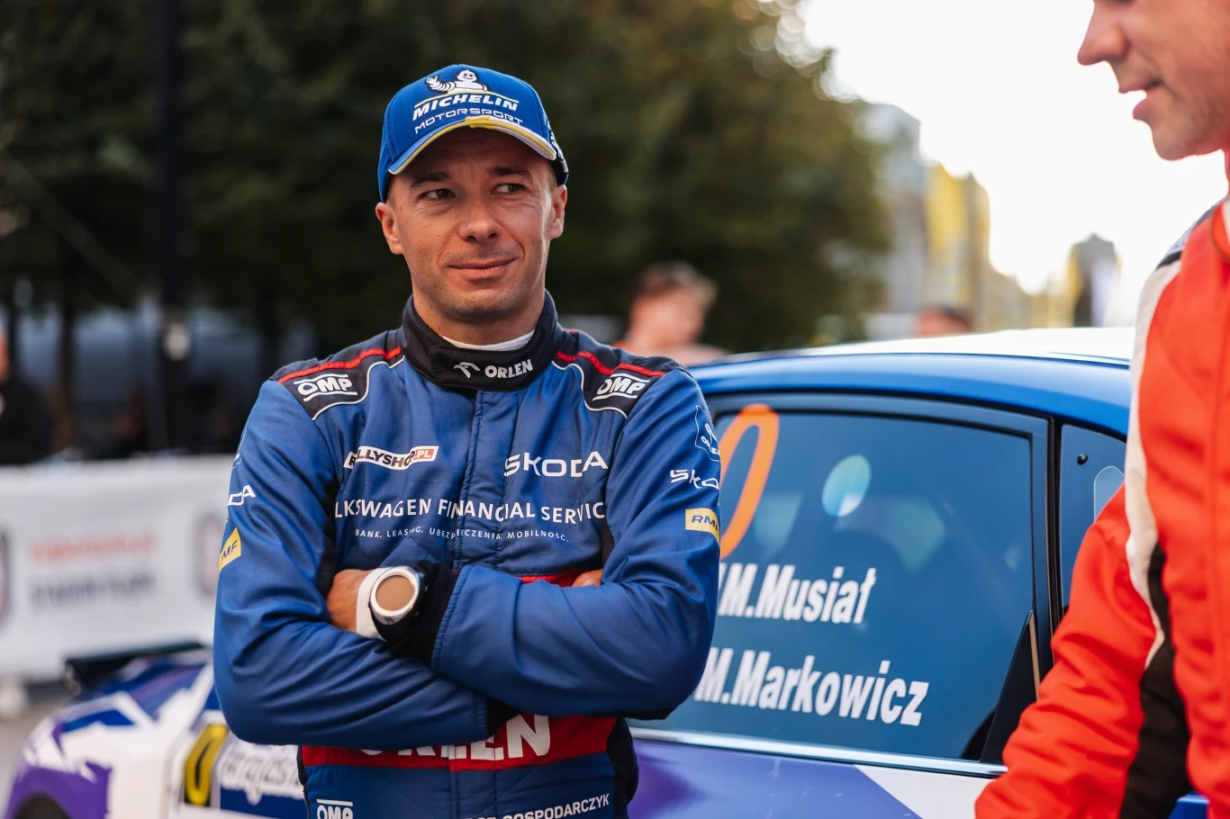 Szymon Gospodarczyk Kierowca rajdowy w kombinezonie i czapce, stojący przy samochodzie wyścigowym z widocznymi oznaczeniami sponsorskimi i nazwiskami na drzwiach, w tle niewyraźne sylwetki ludzi oraz drzewa.