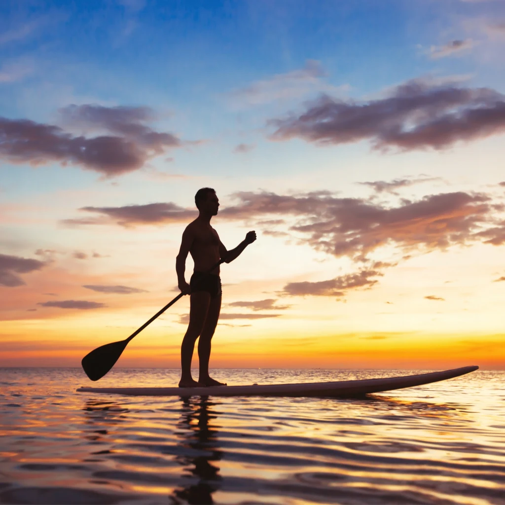 Deski do pływania SUP pozwalają na różnorodne formy aktywności – od rekreacyjnych rejsów, przez ćwiczenia równowagi i fitness na wodzie, aż po wycieczki z bagażem Sylwetka mężczyzny stojącego na desce paddleboard na spokojnej wodzie podczas kolorowego zachodu słońca, z odbiciem światła na powierzchni wody i rozproszonymi chmurami na niebie.