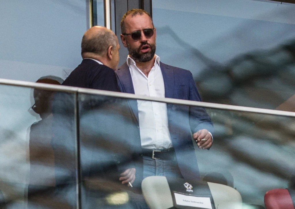 Dwóch mężczyzn w eleganckich garniturach prowadzi rozmowę na trybunach vip sportowego stadionu, jeden z nich ma okulary przeciwsłoneczne i zarost, w tle widoczne przeszklone barierki i puste krzesła.