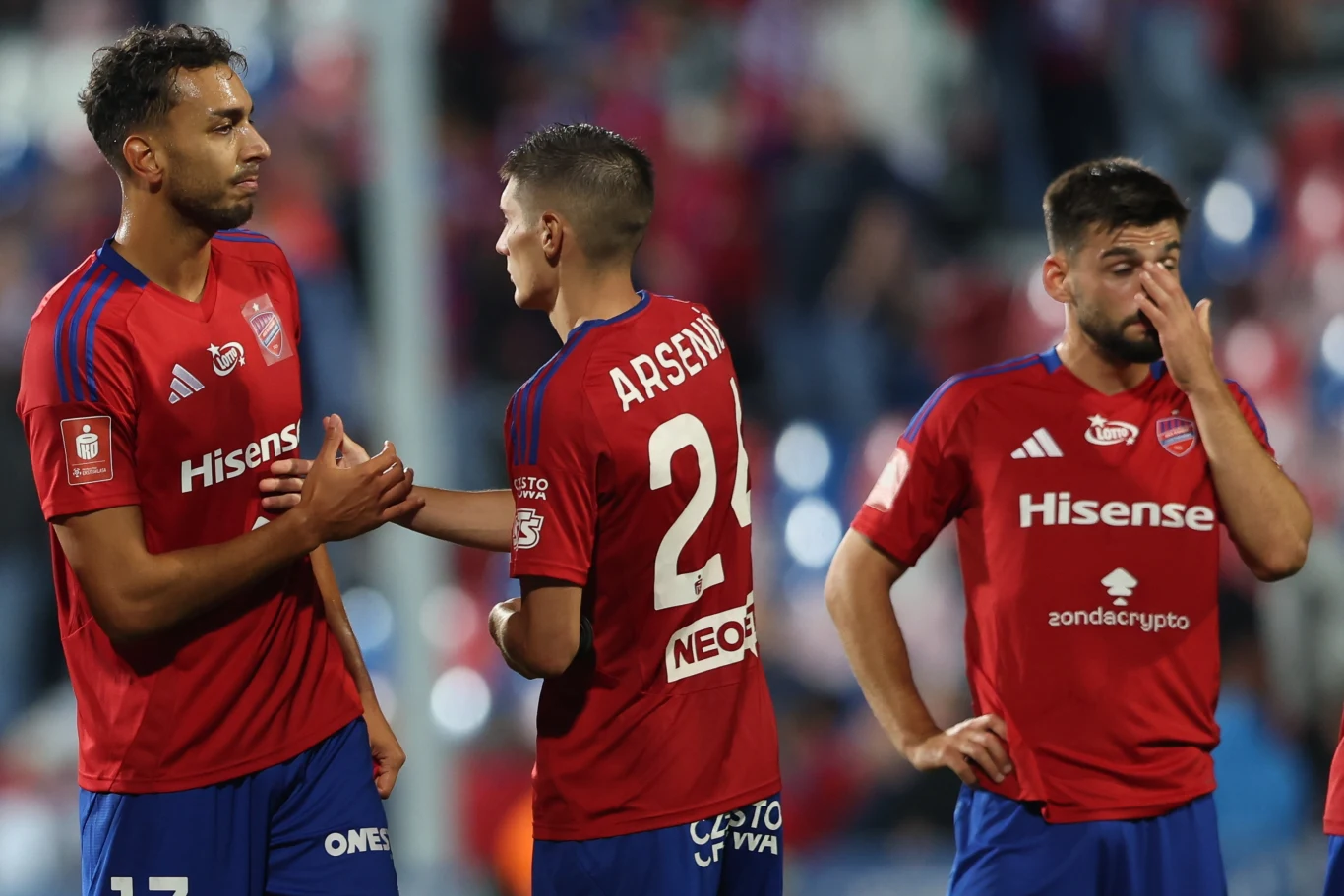Raków Częstochowa - Maccabi Hajfa Trzech piłkarzy w czerwonych koszulkach drużyny Hisense, dwaj z nich rozmawiają lub przekazują sobie gest dłoni, jeden zawodnik dotyka nosa i wygląda na zamyślonego lub rozczarowanego, w tle widoczne zamazane sylwetki kibiców na stadionie