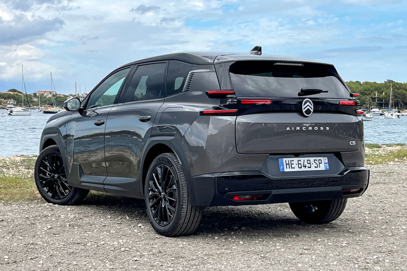 Ostre przetłoczenia, nowe logo i charakterystyczne światła LED. Po tym poznasz francuską nowość. Szary SUV Citroën C5 Aircross zaparkowany na żwirowym terenie przy brzegu jeziora, w tle zacumowane łodzie żaglowe i zielone drzewa.