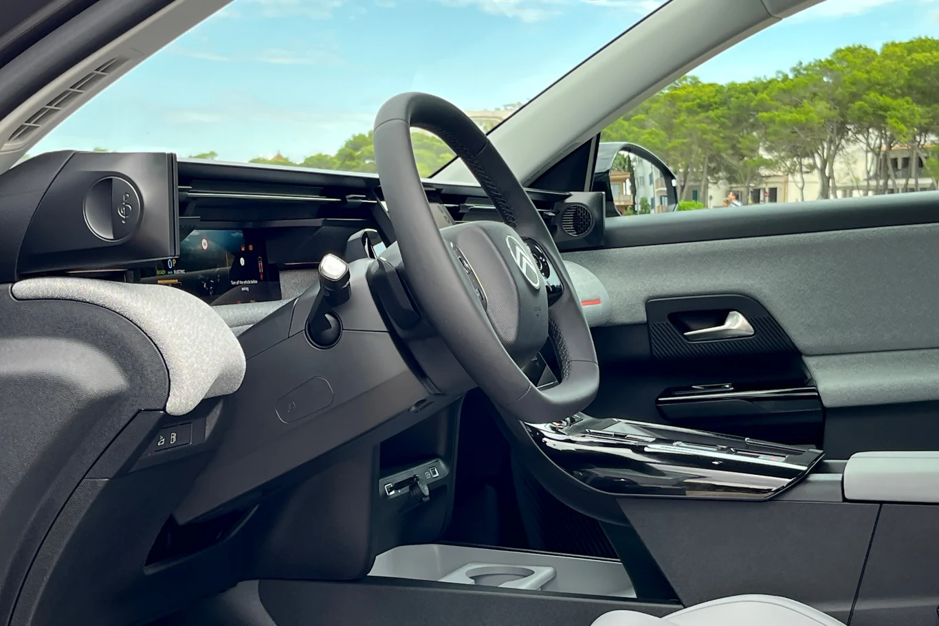 Wnętrze nowego C5 Aircross to udane połączenie komfortu i nowoczesnych technologii. Nowoczesna kabina samochodu elektrycznego z panoramicznym widokiem na deskę rozdzielczą, kierownicą i panelami sterowania, po lewej widoczny ekran, za szybą drzewa i niebo.