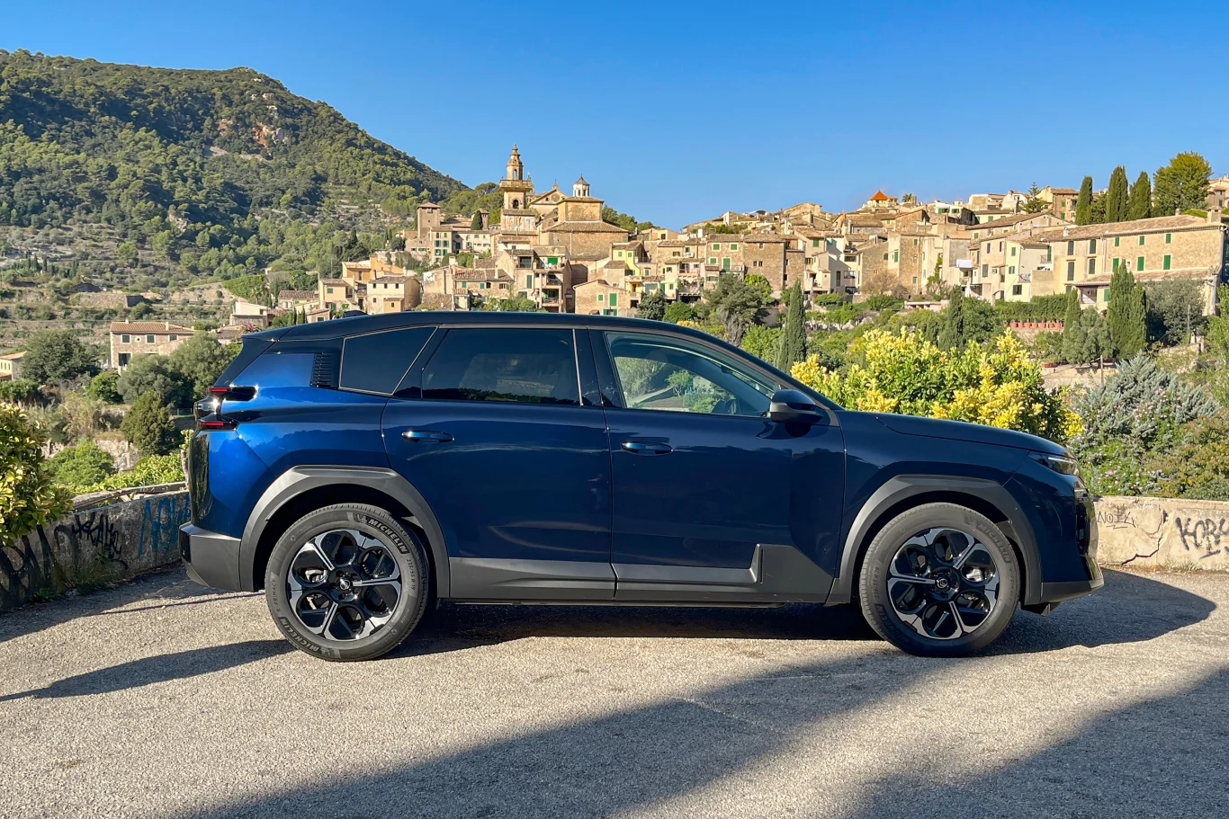 Najważniejszą zmianą w nowym Citroenie C5 Aircross jest jego gama napędowa. Niebieski samochód typu SUV zaparkowany na tle malowniczej miejscowości z kamiennymi domami i otaczającymi ją zielonymi wzgórzami pod błękitnym niebem.