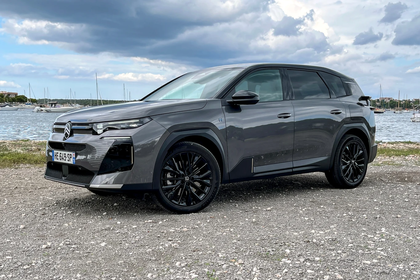 Zamiast diesla do oferty C5 Aircross trafia wariant hybrydowy plug-in oraz elektryczny. Szary nowoczesny SUV stoi na żwirowym podłożu nad wodą z widokiem na przystań i łodzie, na tle pochmurnego nieba. Samochód posiada czarne felgi oraz wyraziste linie nadwozia.