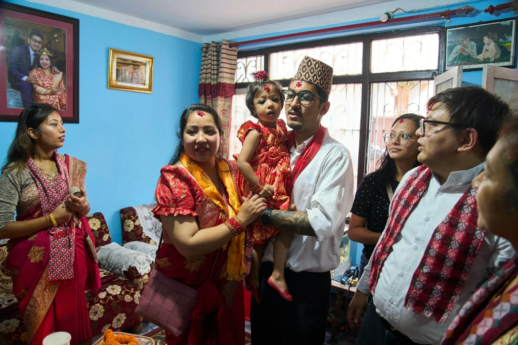 Rodzina ubrana w tradycyjne stroje nepalskie zgromadzona w salonie, z centralnie ustawioną kobietą trzymającą dziecko, wokół kilka innych osób uczestniczących w uroczystości, tło stanowi pokój z niebieskimi ścianami i portretami na ścianach.