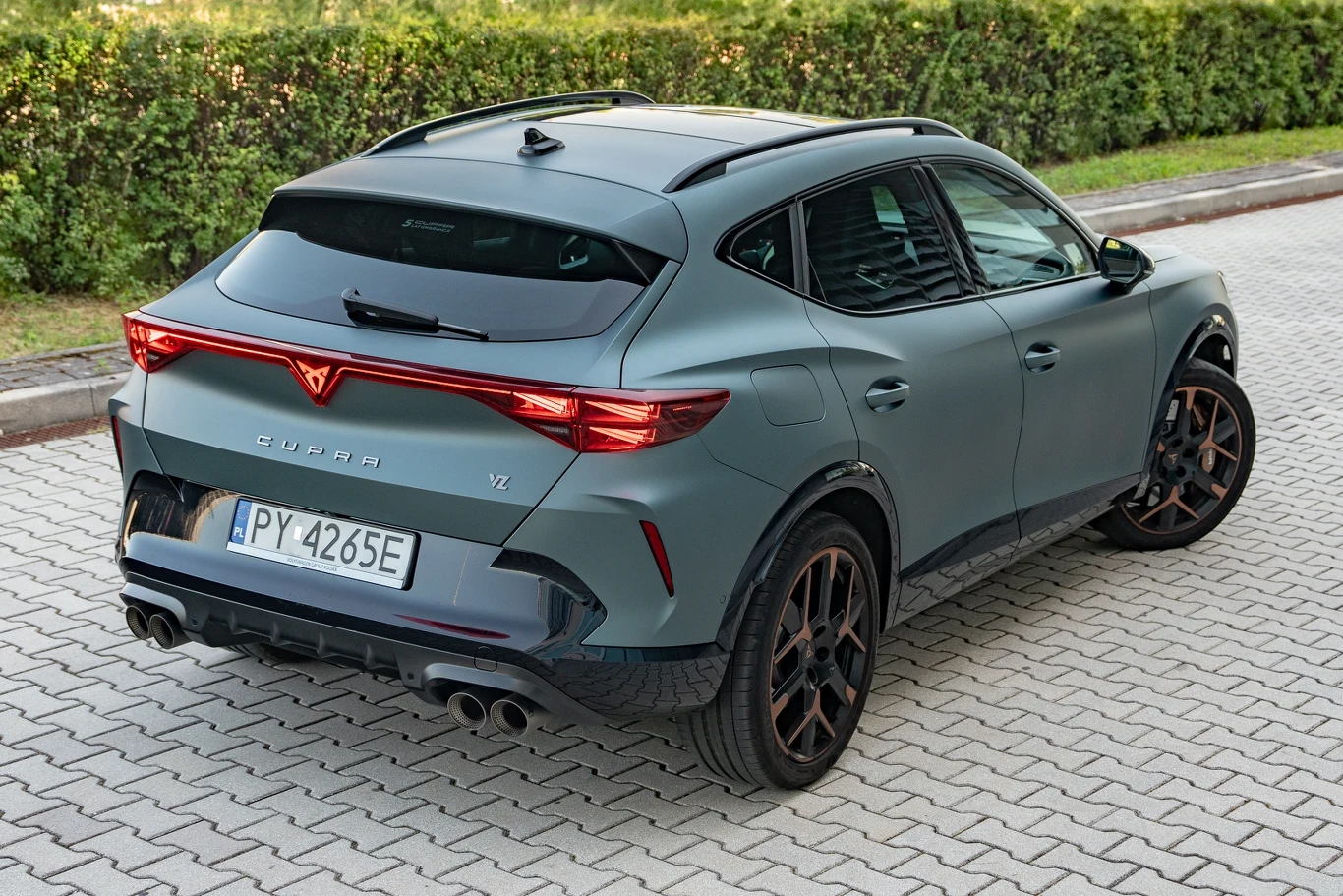 Nowoczesny samochód typu SUV w matowym zielonym kolorze, widok od tyłu, charakterystyczne światła LED, podwójny wydech, czarne felgi, polska tablica rejestracyjna, auto zaparkowane na kostce brukowej obok zielonego żywopłotu.