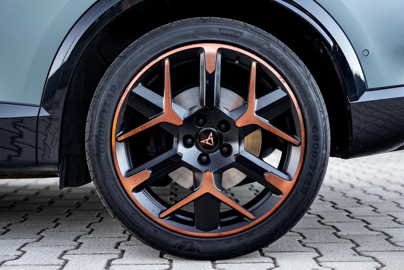 Nowoczesna felga aluminiowa w czarno-miedzianym kolorze z wyraźnymi detalami, zamontowana na oponie, fragment karoserii samochodu oraz kostka brukowa w tle.