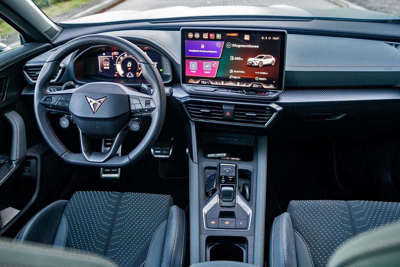 Wnętrze nowoczesnego samochodu z widocznym kierownicą, cyfrowym zestawem wskaźników oraz dużym ekranem dotykowym na desce rozdzielczej, siedzenia pokryte czarną tapicerką z delikatnym wzorem.