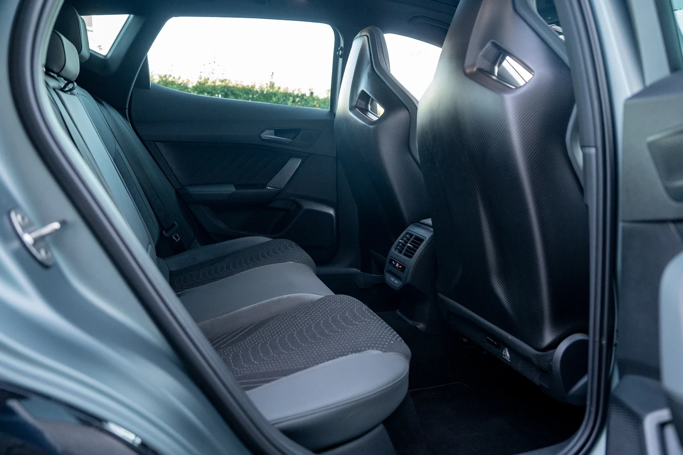 Tylne siedzenia samochodu z nowoczesną tapicerką, środkowym panelem z nawiewami i przestronną przestrzenią na nogi, jasne światło wpadające przez okno.