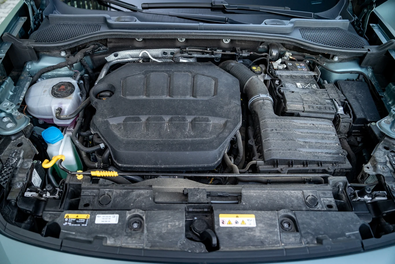 Silnik samochodowy z widocznymi elementami takimi jak zbiornik płynu chłodniczego, filtr powietrza, akumulator oraz różne przewody i pokrywa silnika umieszczona centralnie.