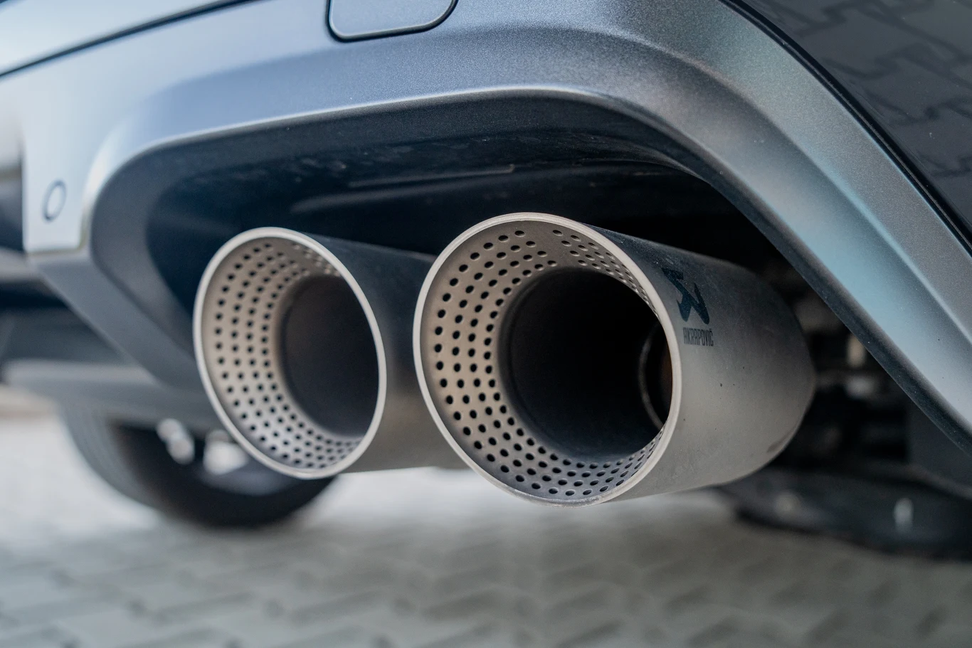 Dwie chromowane końcówki układu wydechowego samochodu wystają spod tylnego zderzaka, powierzchnia wokół wykonana z matowego metalu i plastiku, tło stanowi jasna kostka brukowa