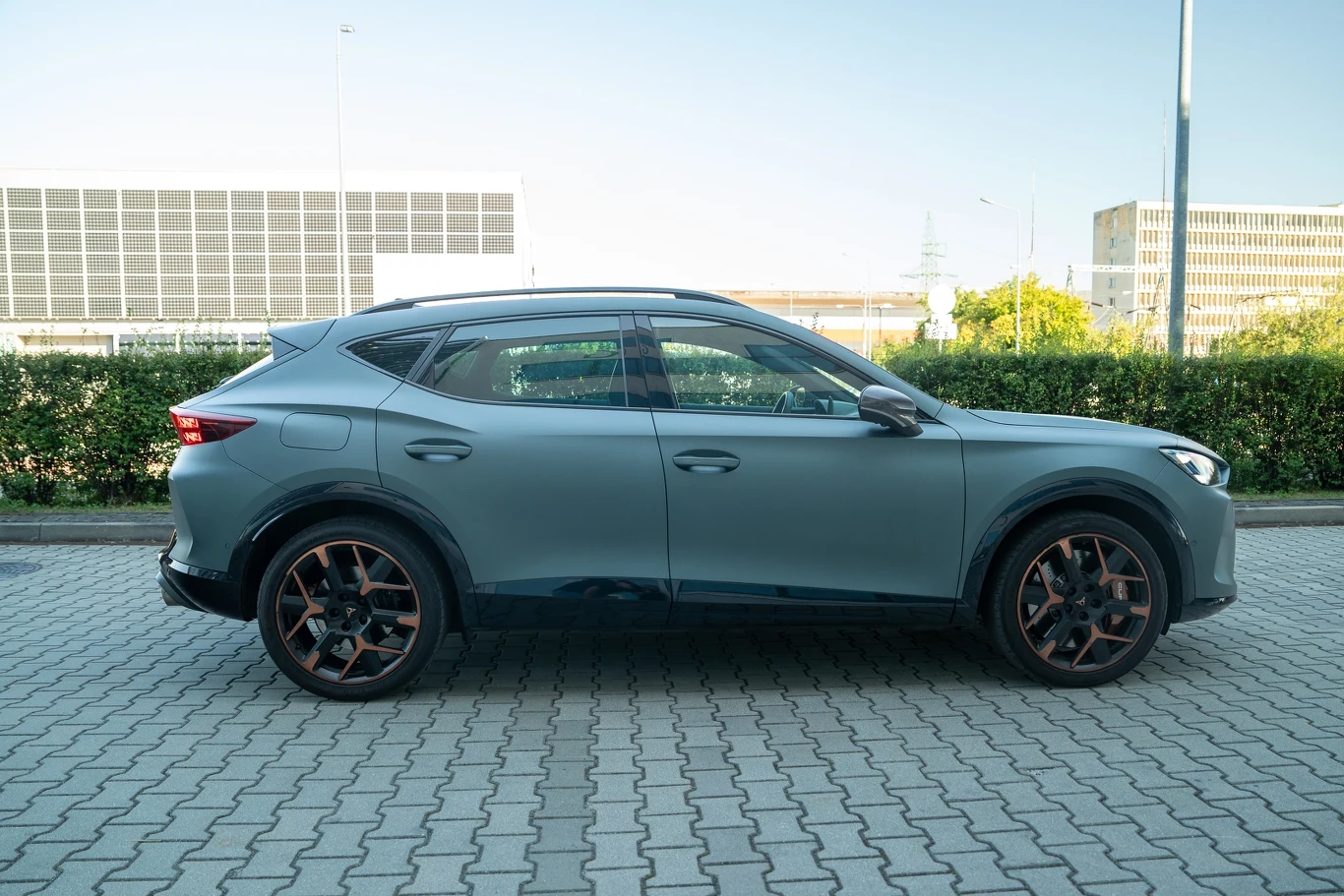 Szary nowoczesny SUV zaparkowany na brukowanym parkingu, widok z boku na tle zielonego żywopłotu i nowoczesnych budynków w oddali.