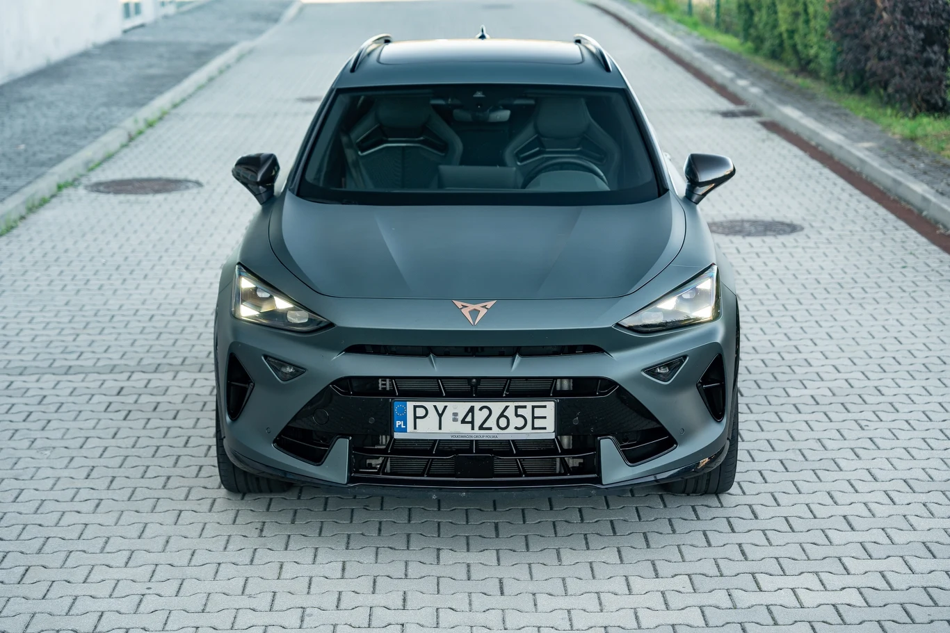 Nowoczesny, sportowy samochód marki Cupra w matowym, grafitowym kolorze zaparkowany na brukowanej ulicy, widok z przodu podkreślający agresywną stylistykę świateł LED i masywny grill.
