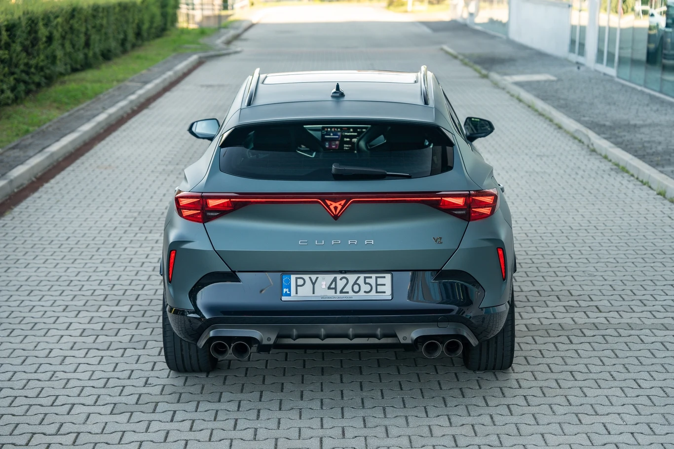 Szary sportowy samochód marki Cupra, widok od tyłu, na tablicy rejestracyjnej oznaczenie PY 4265E, pojazd stoi na kostce brukowej przy zielonym żywopłocie i nowoczesnym budynku.
