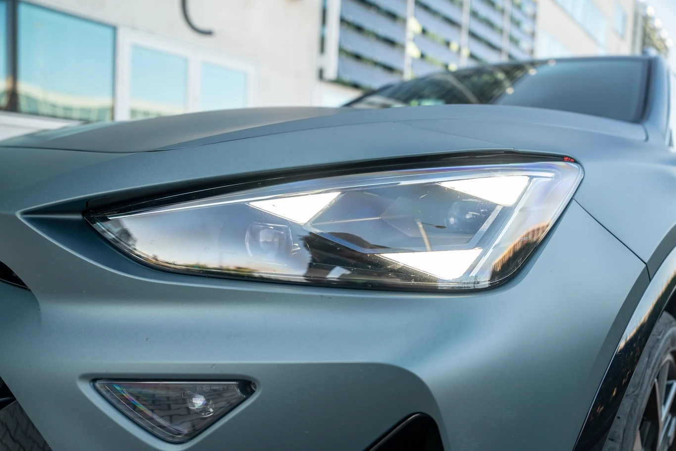 Reflektor nowoczesnego samochodu osobowego w metalicznym niebieskim kolorze z wyraźnie widocznym fragmentem nadwozia oraz liniami maski, w tle widoczne odbicia budynków w szybach.