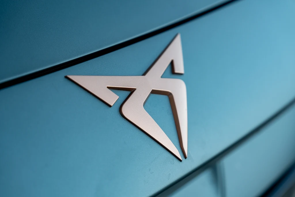 Metaliczne logo Cupra umieszczone na niebieskim karoserii samochodu, wyróżniające się nowoczesnym, geometrycznym kształtem.