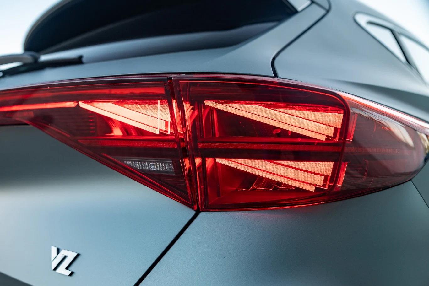 Zbliżenie na tylne światła samochodu z wyraźnie zaznaczonymi czerwonymi kloszami oraz fragmentem srebrnej karoserii i oznaczeniem modelu VZ.