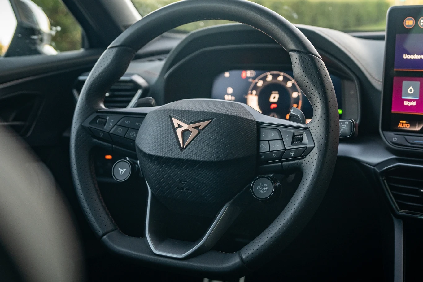 Kierownica sportowego samochodu marki Cupra z dobrze widocznym logotypem na środku, cyfrowy wyświetlacz z prędkościomierzem w tle oraz fragment konsoli środkowej z ekranem dotykowym po prawej stronie.