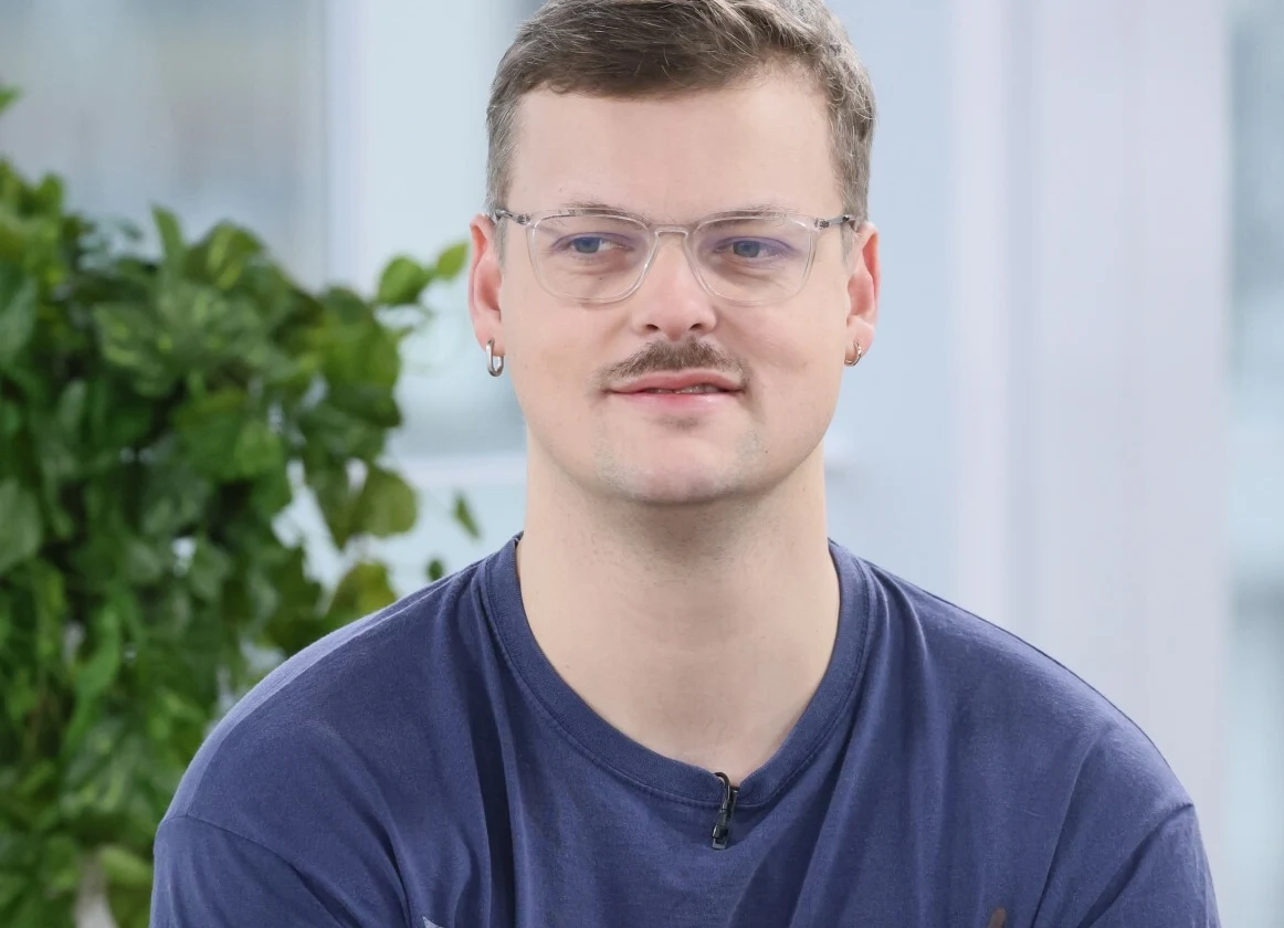 Błażej Król w wywiadzie z PAP Life powiedział o swoim podejściu do posiadania dzieci Mężczyzna w okularach z krótkimi włosami i wąsem w niebieskiej koszulce, patrzący przed siebie, w tle zielona roślina i jasne wnętrze.