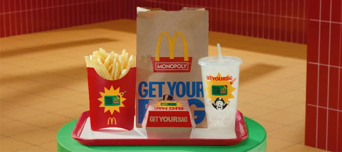 Zestaw fast food składający się z frytek, burgera w opakowaniu oraz napoju w przezroczystym kubku, podanych na tacy obok papierowej torby z logo McDonald’s i nagłówkiem Monopoly.