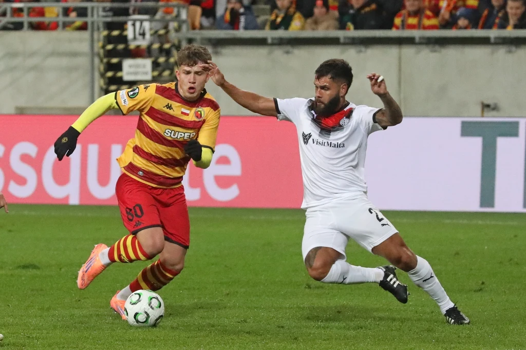 Dwóch piłkarzy walczących o piłkę podczas meczu, jeden ubrany w żółto-czerwony strój klubowy, a drugi w biały komplet, w tle widownia oraz fragment stadionu.