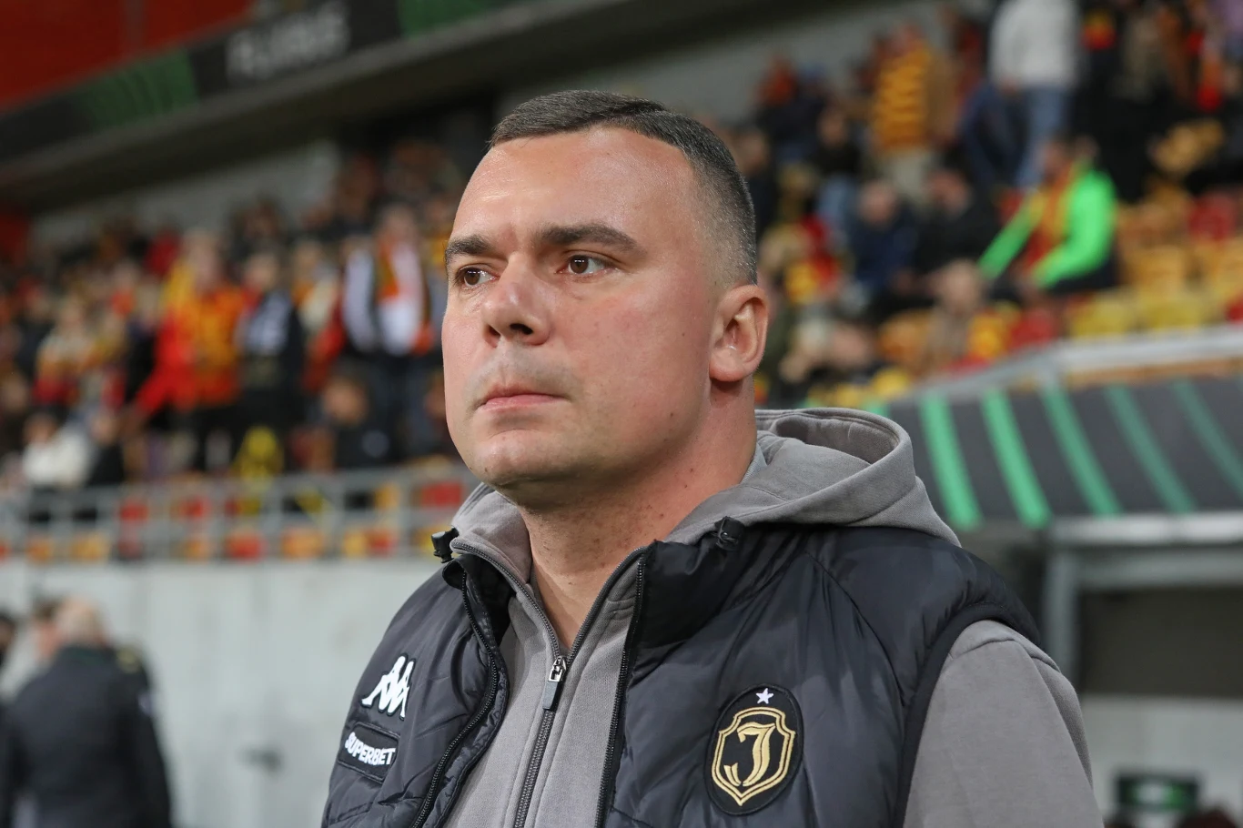 Trener Adrian Siemieniec Mężczyzna w sportowej kamizelce z herbem piłkarskim patrzący przed siebie, w tle trybuny z kibicami ubrane w żółto-czerwone stroje.