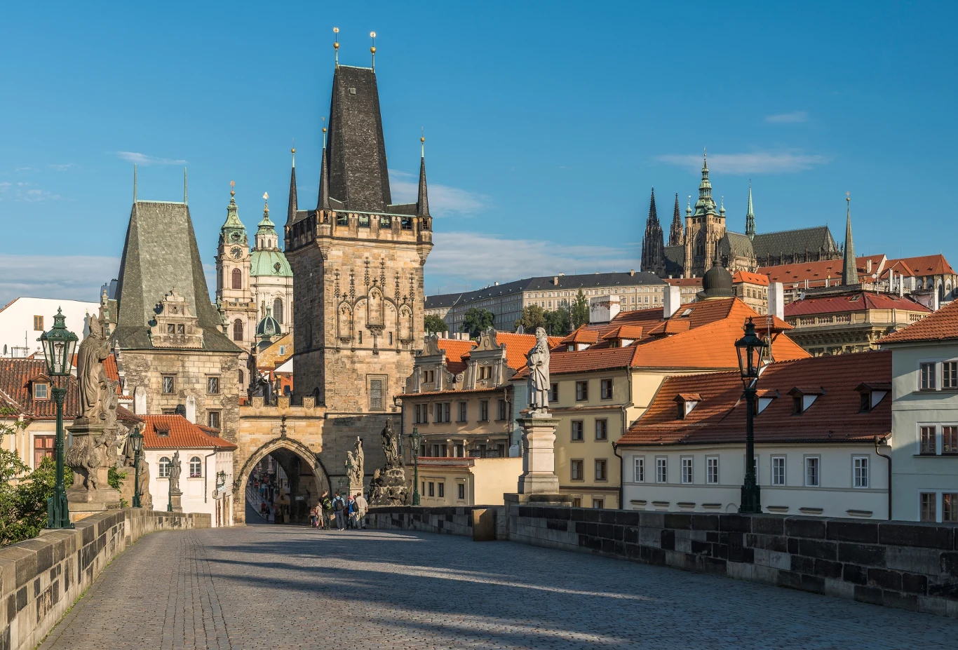 Kamienny most z historycznymi wieżami i rzeźbami, w tle panorama czerwonych dachów i wież zabytkowych budynków Pragi, na horyzoncie kompleks zamkowy z katedrą.