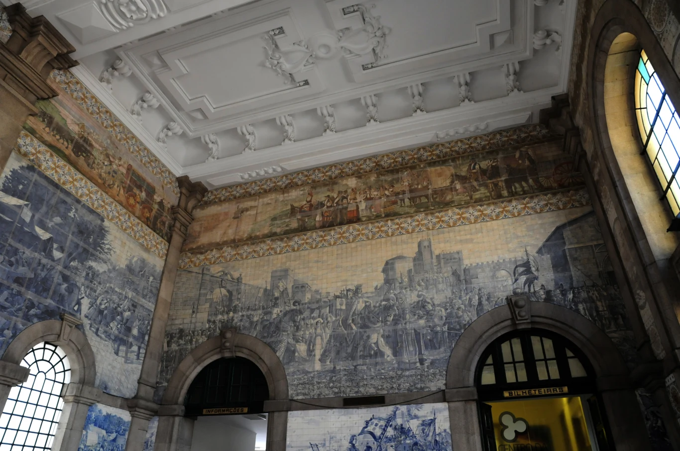 Monumentalne malowidła ścienne i mozaiki przedstawiające sceny historyczne oraz architekturę w zabytkowym wnętrzu budynku, z dużymi łukowatymi oknami i ozdobnym sufitem.