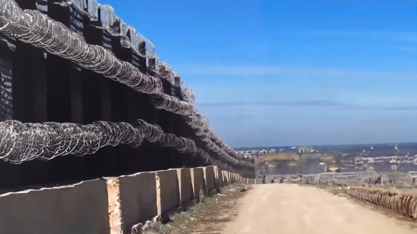 Bariera na granicy z Białorusią to pikuś. Egipt pokazał, jak to się robi Solidna bariera graniczna z wieloma warstwami drutu kolczastego, ciągnąca się wzdłuż żwirowej drogi, w tle rozciąga się rozległy krajobraz z zabudowaniami i równinami.