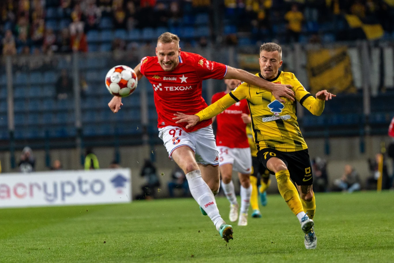 Derby Krakowa zakończyły się remisem Dwóch piłkarzy walczy o piłkę podczas meczu piłkarskiego, jeden ubrany w czerwony strój, drugi w żółto-czarny strój, widoczne dynamiczne starcie na boisku, na dalszym planie trybuny z kibicami.