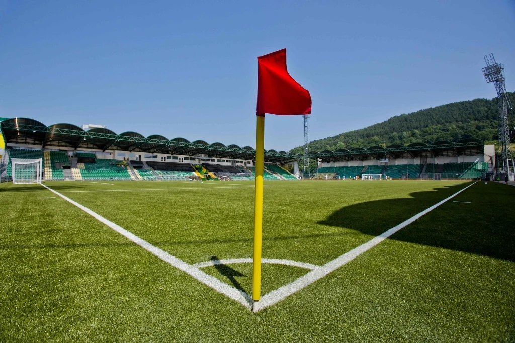 Czerwono-żółta chorągiewka narożna na zielonym boisku piłkarskim, trybuny na tle nieba i wzgórz, stadion pusty, dobrze zadbana murawa.