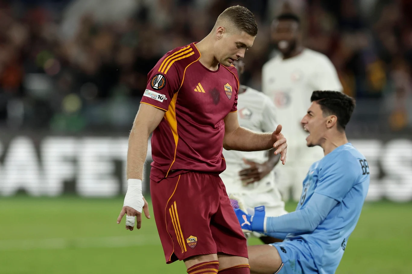Piłkarz w bordowym stroju drużyny AS Roma stoi obok siedzącego na murawie bramkarza w niebieskim stroju, obaj zawodnicy wyglądają na skoncentrowanych, w tle rozmyte sylwetki innych piłkarzy.