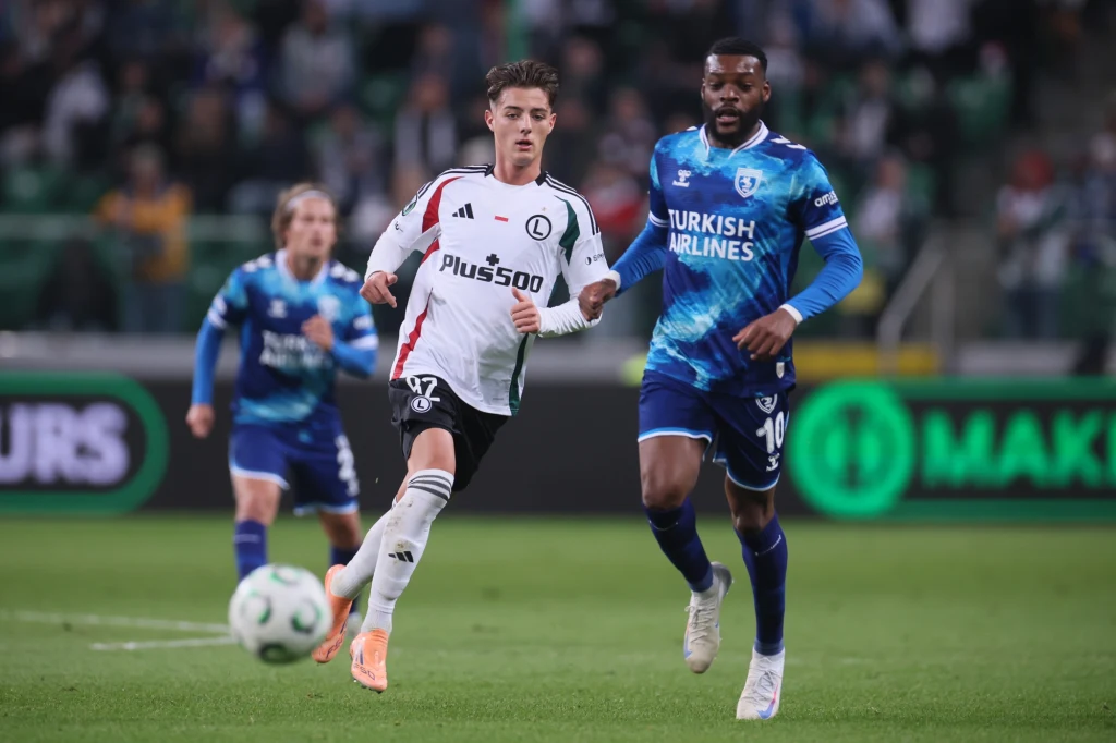 Legia Warszawa - Samsunspor Dwóch piłkarzy biegnących za piłką podczas meczu piłki nożnej na stadionie, jeden ubrany w biały strój z logiem Plus500, drugi w niebieskiej koszulce z logiem Turkish Airlines, rozmyte sylwetki innych zawodników i kibiców w tle.