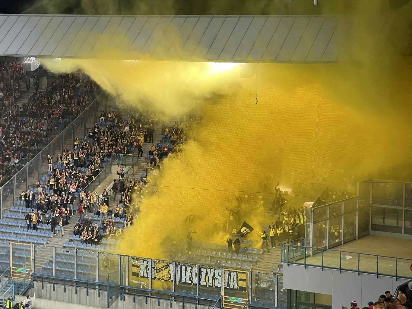 Tłum kibiców piłkarskich na trybunach stadionu otoczony gęstym, żółtym dymem z rac, wiele osób w żółto-czarnych barwach klubowych, widoczny transparent i sektor z aktywnymi fanami.