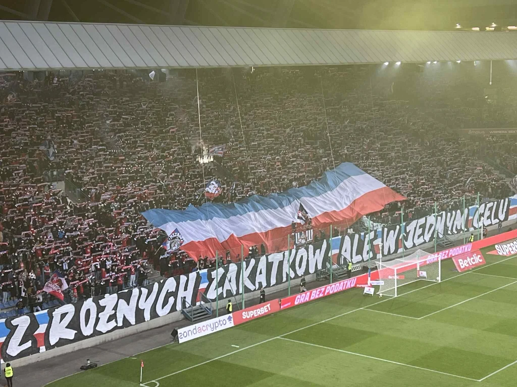 Kibice Wisły Kraków podczas meczu z Wieczystą Tłum kibiców na stadionie tworzy efektowną oprawę, podnosząc dużą flagę w barwach klubowych i trzymając transparent z napisem, który rozciąga się przez całą trybunę, w tle widoczne są trybuny i boisko piłkarskie.