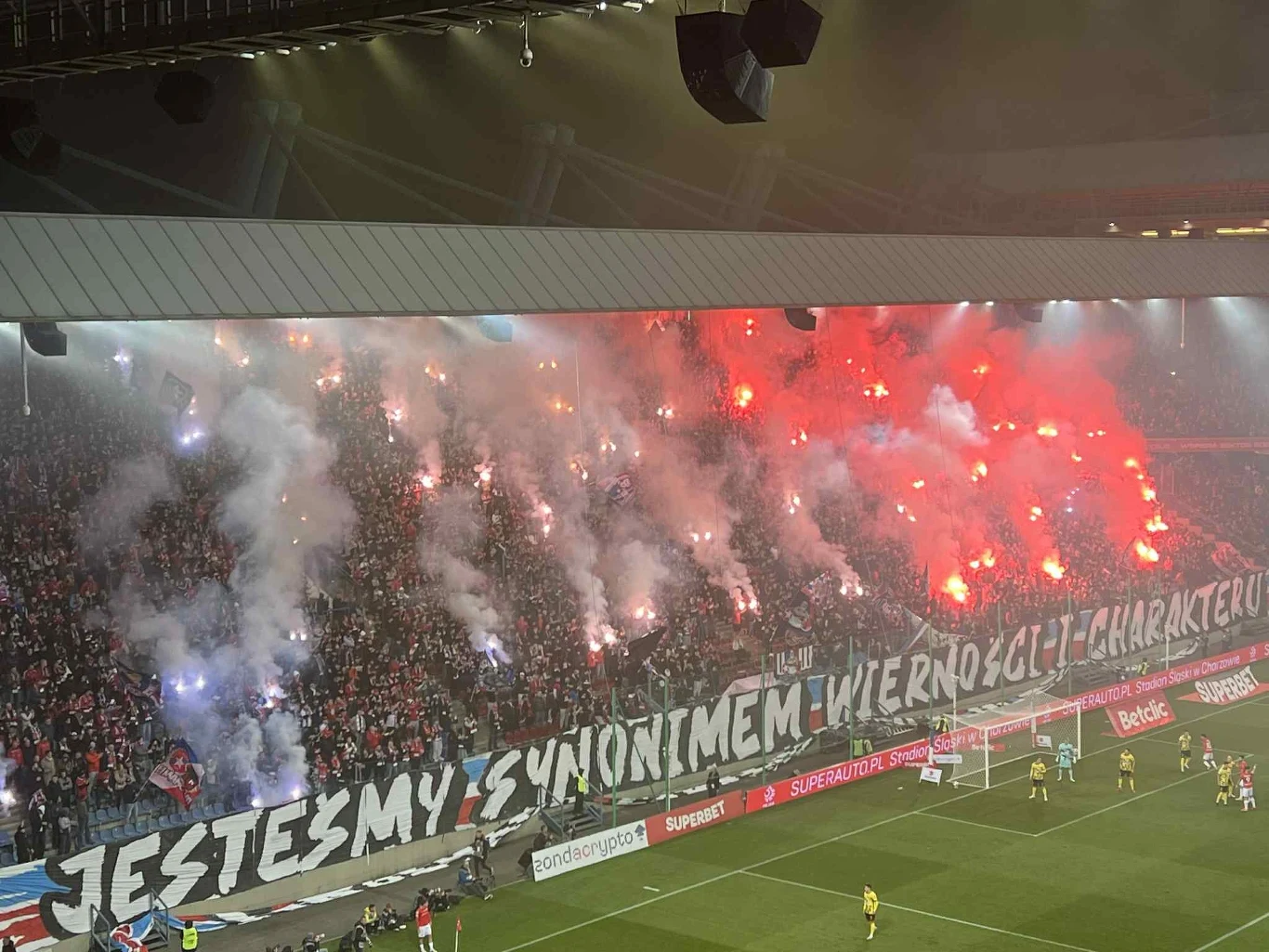 Tłum kibiców na trybunach stadionu piłkarskiego odpala czerwone race i trzyma duży transparent z napisem 'JESTEŚMY SYNONIMEM WIERNOŚCI I CHARAKTERU', wokół panuje gęsty dym i atmosfera emocji sportowych