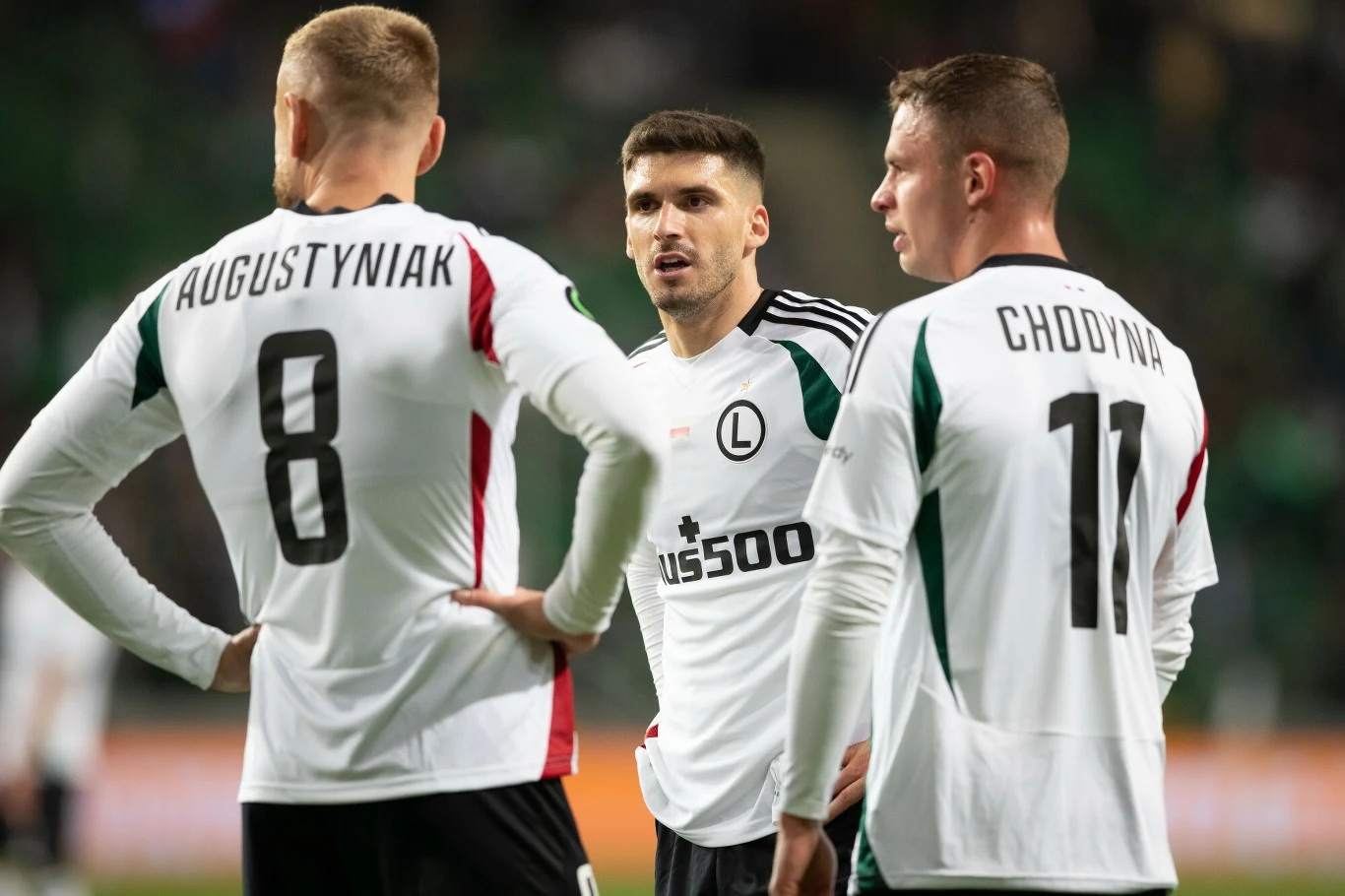 Legia Warszawa - Samsunspor Trzech piłkarzy Legii Warszawa w białych strojach drużyny podczas dyskusji na murawie, w tle rozmyta trybuna stadionu.