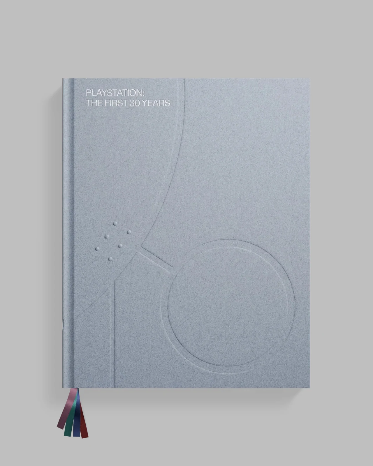 Album "PlayStation: The First 30 Years" w standardowym wydaniu Szara okładka książki z napisem 'PlayStation The First 30 Years', tłoczonymi geometrycznymi elementami oraz kolorowymi tasiemkami indeksującymi wystającymi z dolnej części książki.