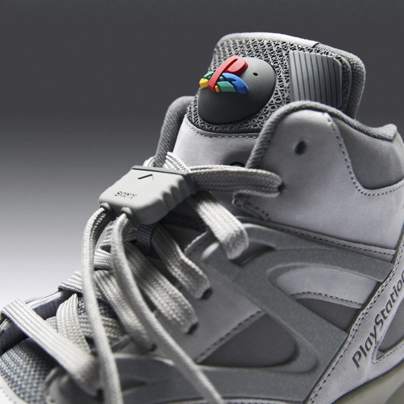 Pump Omni Zone II z kolekcji Reebok x PlayStation But sportowy w odcieniach szarości z charakterystycznym elementem PlayStation na języku, zawiązany grubą, szarą sznurówką, logo PlayStation w kolorze kolorowym oraz napisem na boku buta.