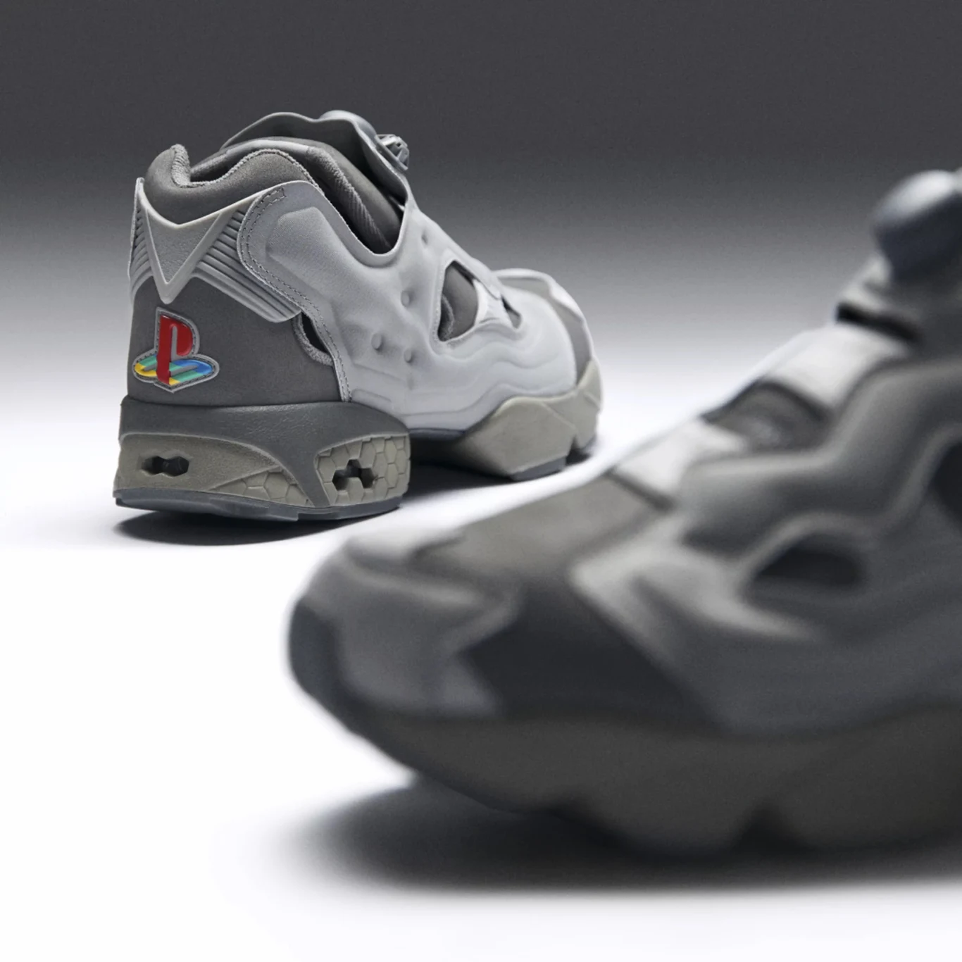 Reebok InstaPump Fury 94 w barwach PlayStation Sportowe buty w odcieniach szarości z wyraźnym, kolorowym logo PlayStation na pięcie, stojące na neutralnym tle, w centrum ostrości jeden but, drugi rozmyty na pierwszym planie.