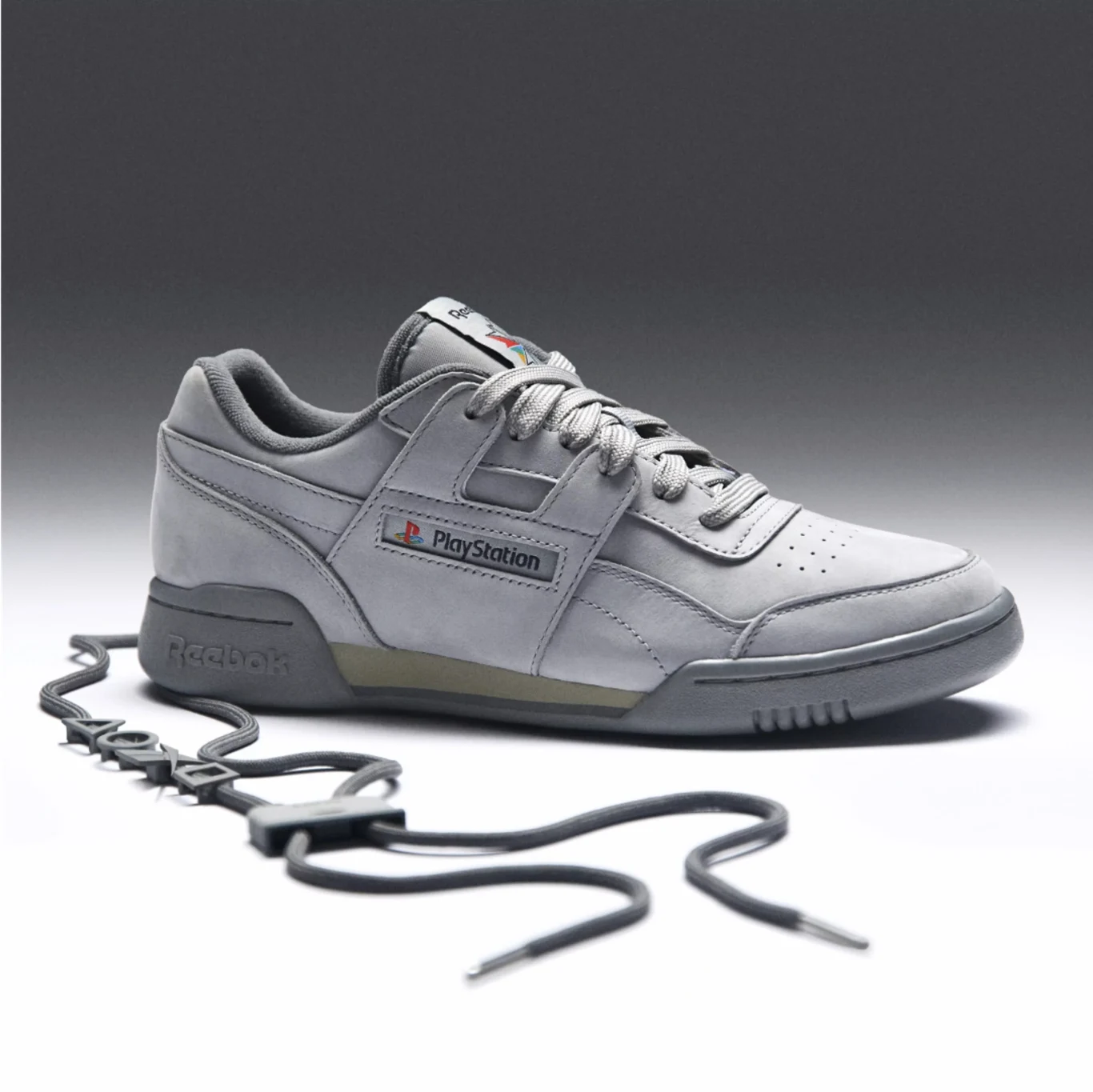 Reebok Workout Plus w wydaniu dla graczy Szary sneaker marki Reebok inspirowany stylistyką PlayStation, z charakterystycznym logo konsoli i rozpoznawalnym napisem, dopełniony czarnymi sznurówkami ułożonymi na powierzchni wokół buta.