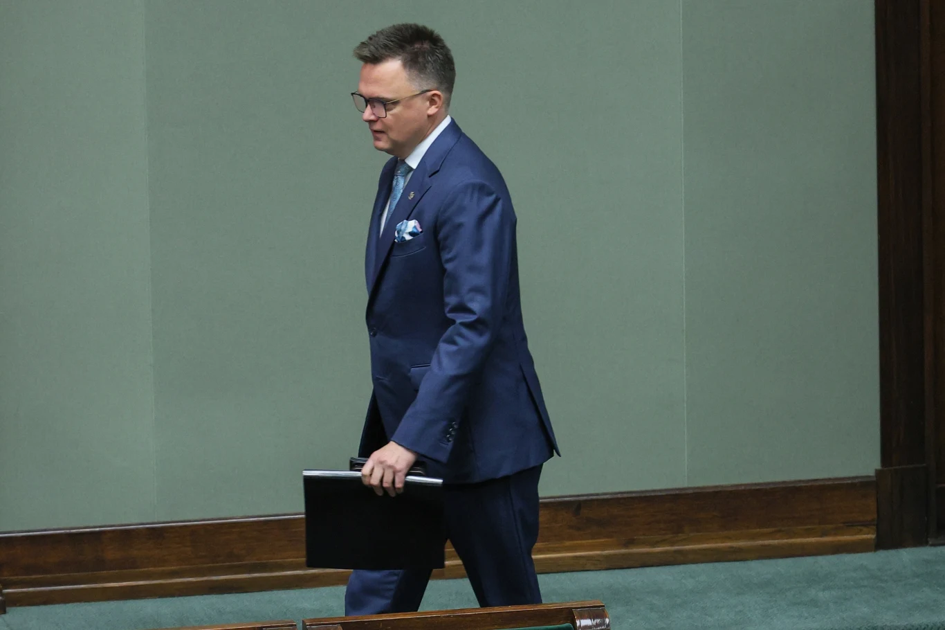 Marszałek Sejmu Szymon Hołownia Mężczyzna w granatowym garniturze i okularach idący przez salę, trzymający czarną teczkę, otoczenie wskazuje na oficjalne wnętrze, prawdopodobnie parlamentarne.