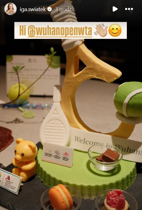 Słodki deser z elementami inspirowanymi tenisem, w tym figurka rakiety tenisowej, piłki tenisowej oraz misia, a także różne kolorowe słodkości i tabliczka powitalna z napisem Welcome to Wuhan.