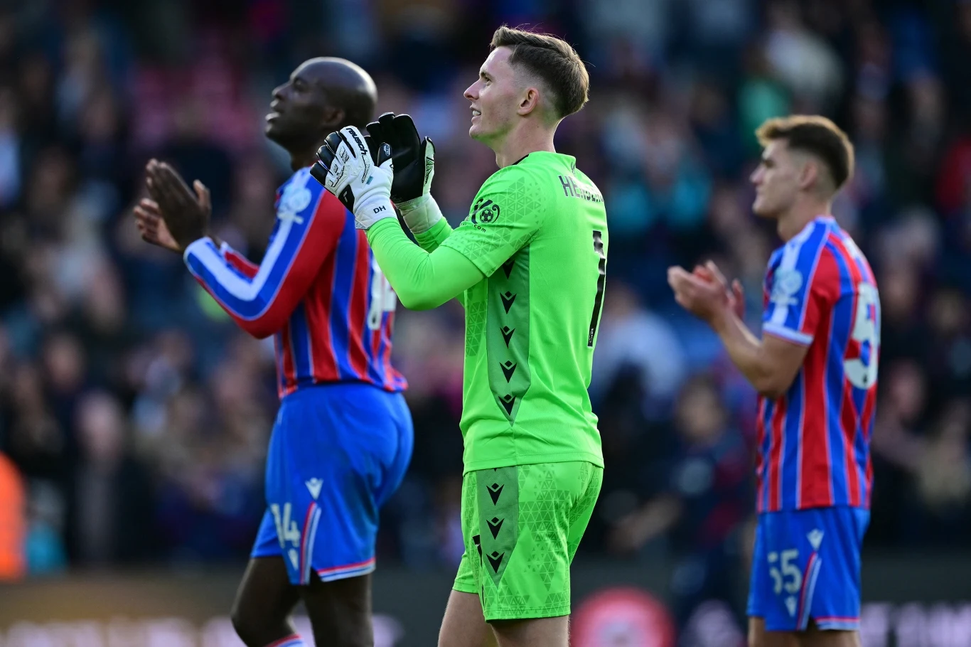 Piłkarze klubu Crystal Palace na boisku w trakcie meczu, bramkarz w neonowym stroju i zawodnicy w czerwono-niebieskich koszulkach biją brawo, rozmyte tło trybun z kibicami.