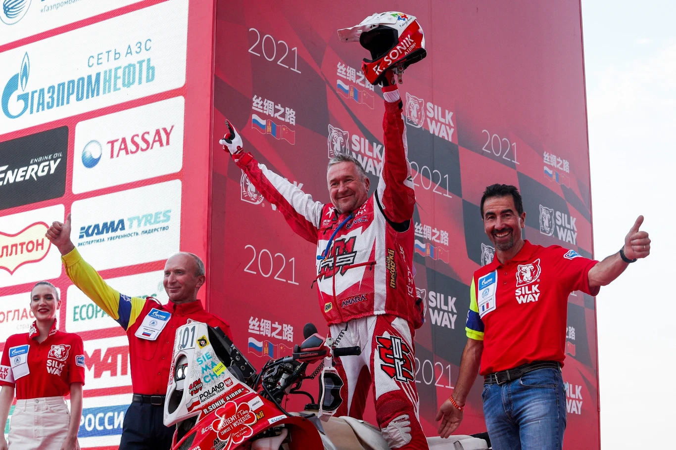 Zwycięzca rajdu motocyklowego stoi na podium z podniesionym kaskiem i uniesionymi rękami w geście radości, otoczony przez członków zespołu i osoby wspierające; w tle widoczne logo sponsorów i czerwone dekoracje z nazwą rajdu Silk Way 2021.